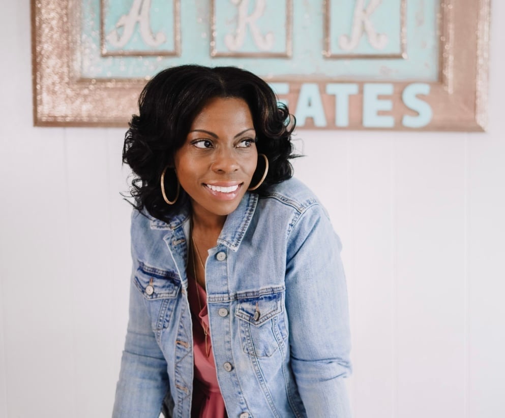 Woman wearing light blue denim jacket and red top, smiling at camera with rustic signage in background